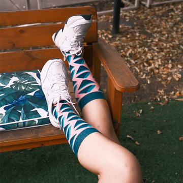 Person relaxing on a wooden bench wearing knee-high socks with geometric pink and teal patterns, paired with white sneakers. The socks add a stylish and playful touch to the casual outdoor setting.