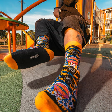 Person sitting outdoors wearing colorful jacquard socks with bold patterns in blue, yellow, and orange. The socks feature unique designs with a playful and vibrant style, complementing a casual outfit in a sunny park setting.